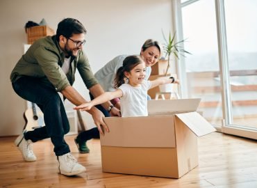 Happy little girl with arms outstretched having fun being pushed by her parents in carton box at new home